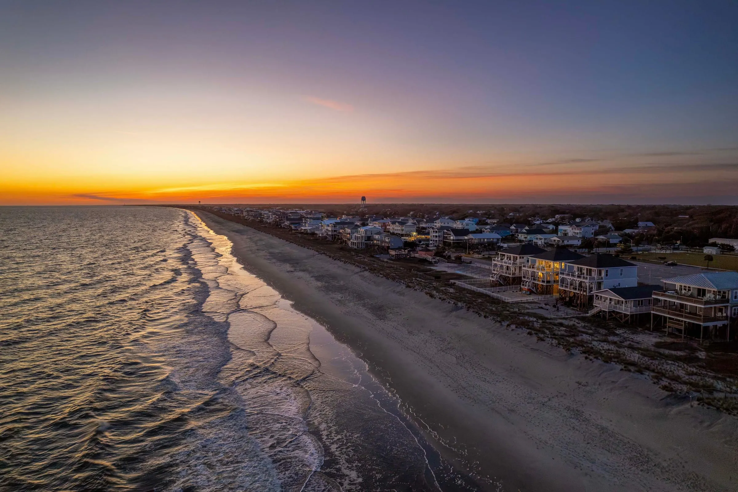 Aerial dusk view of Oak Island: ocean on the left, wet sand running toward the horizon, a row of lit beach houses on the right, and the water tower in the distance